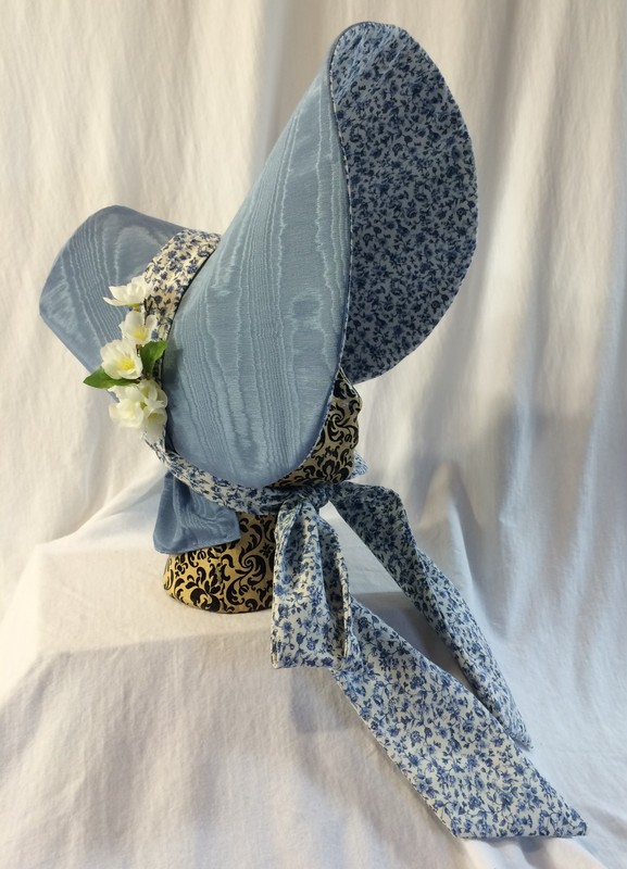 This poke bonnet is for dressier occasion than the bonnet above.  This has a small stiff crown and oversized bill covered with a blue jacquard with a lining and tie made with a flowered cotton print. Flowers adorn the side of the crown.  A black velvet bonnet may have been worn when mourning. 